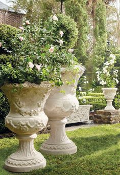 two large white vases with flowers in them on the grass near some bushes and trees