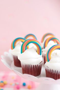 cupcakes with white frosting and rainbow decorations on a cake platter against a pink background