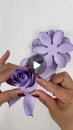 two hands holding purple paper flowers on a white surface with one hand reaching for the flower