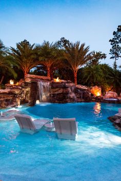 two lawn chairs sitting in the middle of a pool with waterfall and lights on it