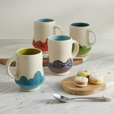 four coffee mugs with different designs on them sitting next to a cutting board and spoon