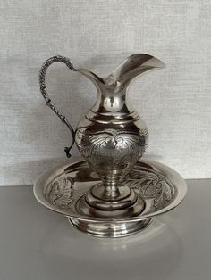 a silver pitcher sitting on top of a metal plate with a chain hanging from the handle