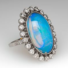 This gorgeous ring is centered with one (1) oval cabochon cut natural crystal opal and set into a bezel setting. The opal is bordered with twenty-two (22), bezel set, round brilliant cut diamonds, The top face of the shank is accented with three (3), bead set, round brilliant cut diamonds on each side. The gemstones are bordered with milgrain edging. The ring measures 29.1mm at the top, rises 10.6mm above the finger, tapering to 1.8mm wide and 1.1mm thick at the base of the shank. This ring is currently a size 6. Fine Jewelry Oval Cabochon Opal Ring, Oval Cabochon Opal Ring Fine Jewelry, Oval Cabochon Opal Ring In Fine Jewelry, Exquisite Oval Multi-stone Opal Ring, Exquisite Multi-stone Oval Opal Ring, Elegant Opal Cabochon Ring, Oval Opal Ring With Bezel Setting For Formal Occasions, Oval Cabochon Diamond Opal Ring, Oval Opal Rings With Bezel Setting