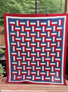 a red, white and blue quilt sitting on top of a wooden table next to a potted plant