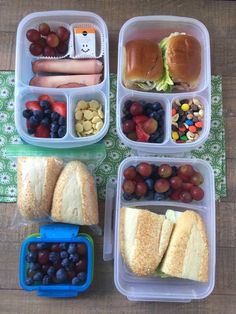 four plastic containers filled with sandwiches, fruit and veggies on top of a wooden table