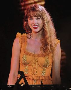 a woman with long blonde hair wearing a yellow dress and smiling at the camera while standing in front of a microphone