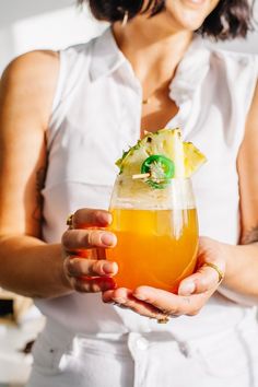 a woman holding a drink with pineapple in it's hand and wearing a white shirt