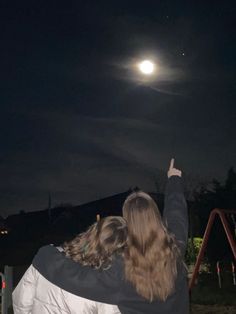 two people looking up at the moon in the night sky with their arms raised to the side