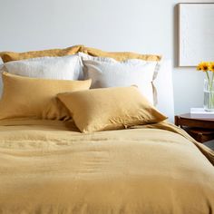 a bed with yellow sheets and pillows in a white room next to a vase filled with sunflowers