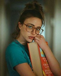 a woman with glasses holding a book in her hand and looking at the camera while leaning on it