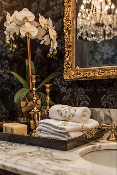 towels are stacked on a tray in front of a sink with a mirror and chandelier