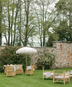 a lawn with chairs and an umbrella in the grass next to a stone wall that is surrounded by trees