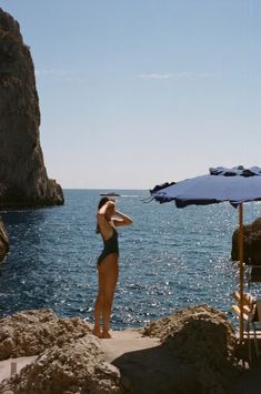 a woman in a bathing suit standing on rocks near the ocean with an umbrella over her head