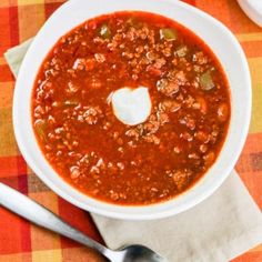 a white bowl filled with chili soup on top of a checkered table cloth next to two silver spoons