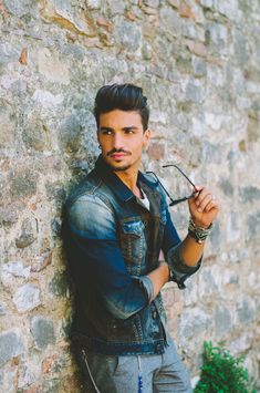 a young man leaning against a stone wall with his hands on the hips and looking at the camera