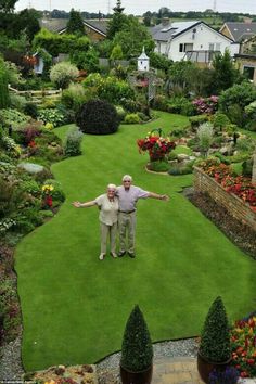 two older men standing in the middle of a lush green yard with lots of flowers