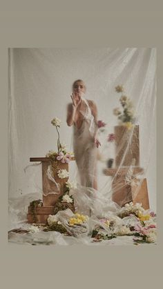 a woman standing in front of a white backdrop with flowers on the floor and behind her is a wooden box