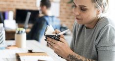 a woman sitting at a table looking at her cell phone while holding a pen and paper