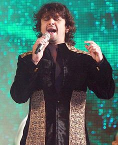 a man holding a microphone in his right hand and wearing a black suit with gold trim