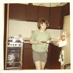 an old photo of a woman holding a plate in her hand while standing in front of the stove