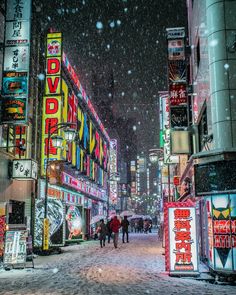 people walking down a snowy street in the middle of a city with neon signs on it