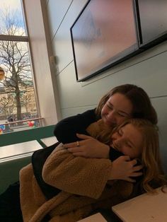 two women hugging each other while sitting at a table in front of a large window