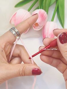 two hands with red nail polish holding pink crochet hooks next to tulips