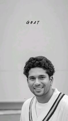 a black and white photo of a man with curly hair smiling at the camera, in front of a sign that says goatt