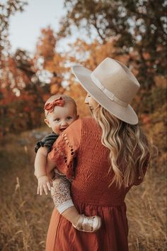 a woman in an orange dress holding a baby wearing a white hat while standing in tall grass