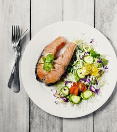 a white plate topped with salmon and salad next to a fork on top of a wooden table