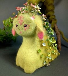 a yellow stuffed animal with flowers on its head sitting next to a branch and green leaves