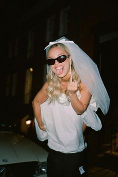 a woman wearing sunglasses and a veil on her head giving the peace sign while standing in front of a car