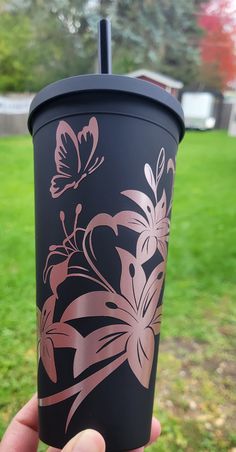 a hand holding up a black and pink cup with flowers on the outside, in front of a grassy area