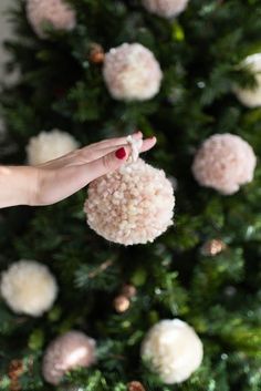 a hand holding a pom - pom ornament in front of a christmas tree