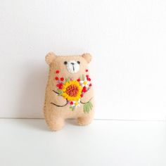 a teddy bear with flowers on its chest is standing next to a white wall and two other stuffed animals are behind it