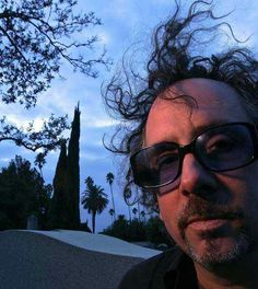 a man with long hair and glasses standing in front of a palm tree at dusk