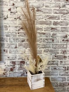 a plant in a white square vase on a wooden table next to a brick wall