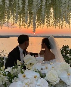 a bride and groom sitting at a table with flowers in front of the sun setting