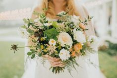 a woman holding a bouquet of white and orange flowers in front of a building with string lights