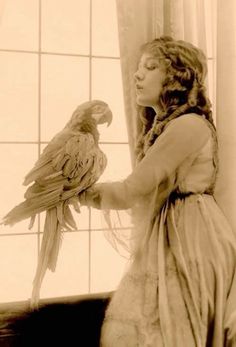 a woman is holding a bird in her hand while standing next to a window with curtains