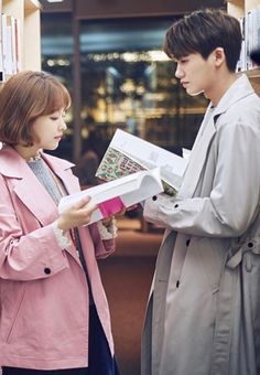 two people standing in front of bookshelves looking at one another and holding an open book