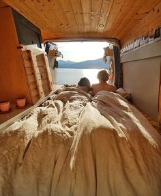 two people are laying in bed looking out the window at the water from inside their camper