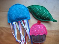 two felt toys are sitting next to each other on a wooden surface, one has a green leaf and the other is a pink cupcake