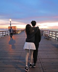 a man and woman standing on a pier looking at the ocean while the sun sets