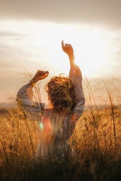 a woman standing in tall grass with her hands up to the sky and arms raised