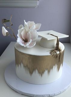 a white and gold graduation cake with flowers on top, sitting on a table in front of a window