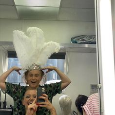 two women taking a selfie in front of a mirror with white feathers on their head