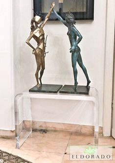 two bronze statues are sitting on a glass table in front of a framed art piece