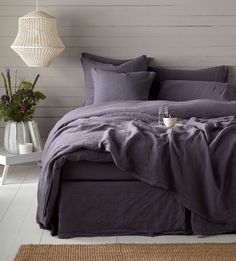 a bed with purple linens and pillows in a white room next to a lamp