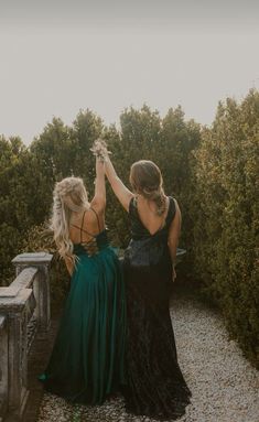 two women in long dresses are standing on a stone wall and touching each other's hands